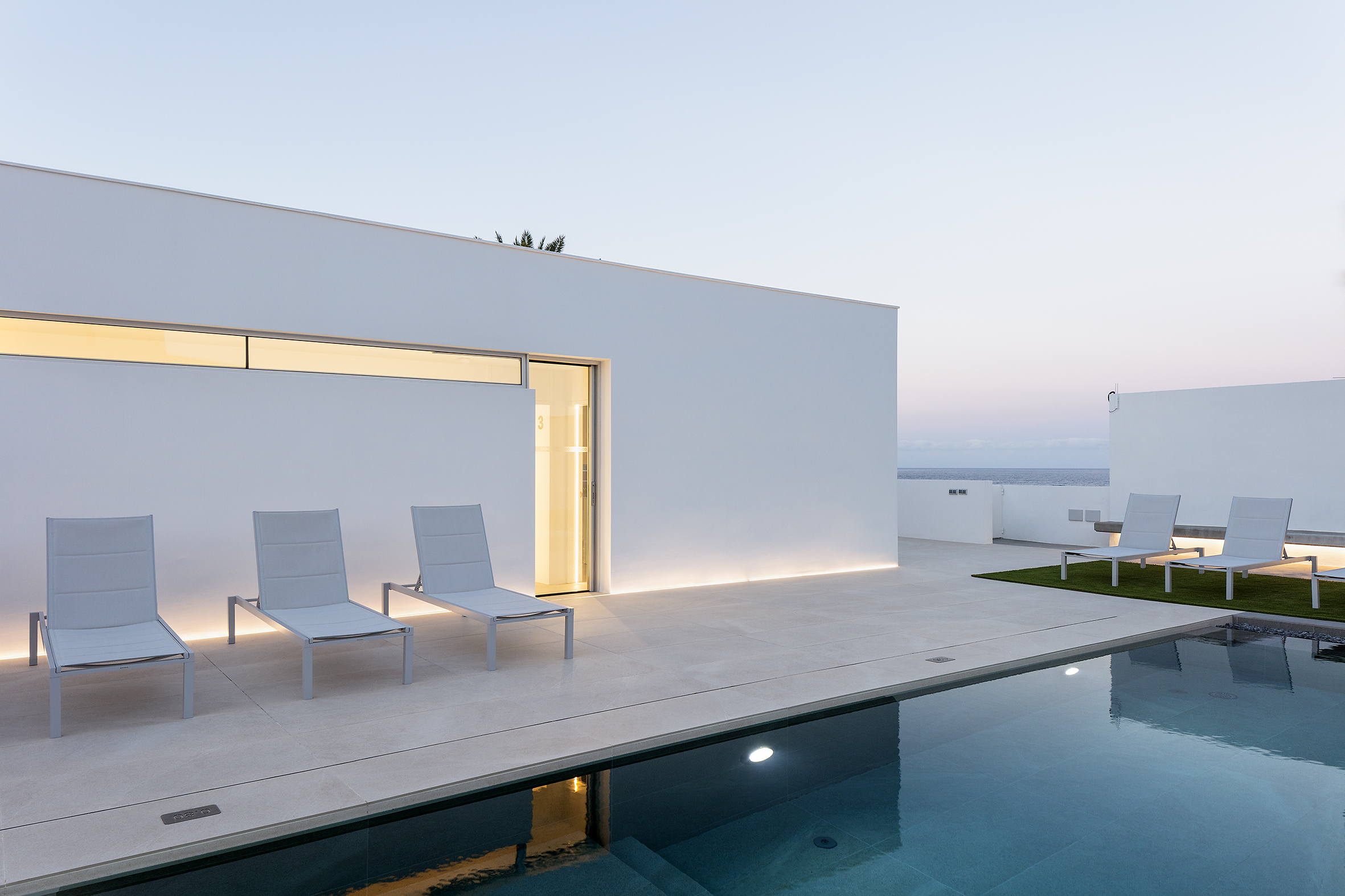 Vista de una terraza moderna con sillas de descanso junto a una piscina, un edificio blanco en el fondo, y un cielo claro al atardecer.